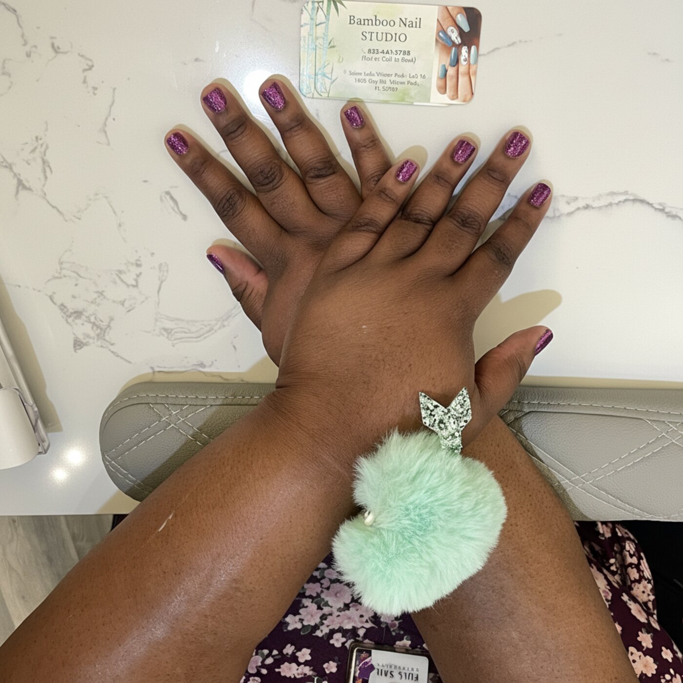 Sparkling purple glitter gel manicure on marble countertop with mint pom-pom